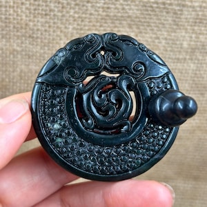May include: Dark green jade pendant with intricate carvings. The circular pendant features a central swirling design and a decorative border of small circles. A small knob extends from the side. The pendant is held in a hand.