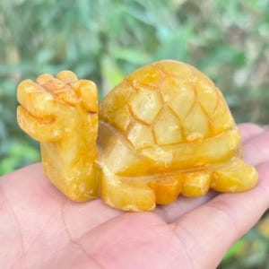 Peut inclure: Une figurine de tortue sculptée, jaune-orangé. La carapace de la tortue présente un motif géométrique, et sa tête et ses pattes sont visibles. La figurine est tenue dans une main, avec un arrière-plan vert flou.