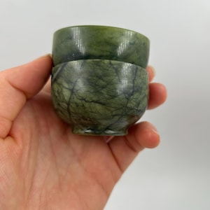 Op de afbeelding: Twee kleine groene stenen kopjes op elkaar gestapeld. De onderste beker heeft een gemarmerd patroon, de bovenste is effen groen. De kopjes worden in een hand gehouden tegen een witte achtergrond.
