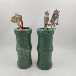 May include: Two green jade-like bamboo-shaped containers. One holds a silver butterfly-shaped object and a red item. The other holds a silver object with a dragon head and a silver chain with bells. The containers are on a white background.
