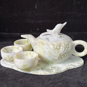 Puede incluir: Un juego de té de piedra tallada con una tetera, tres tazas y una bandeja. La tetera tiene un asa en forma de pájaro y una tapa con una figura de pájaro. El juego es de color verde pálido y blanco.