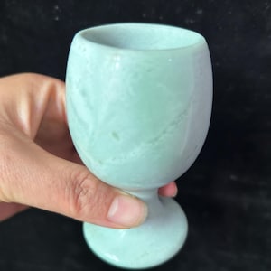 May include: A light green, egg-shaped goblet made of stone. The cup has a rounded base and a short stem. The stone has a marbled appearance with subtle variations in color. The goblet is held by a hand against a dark background.