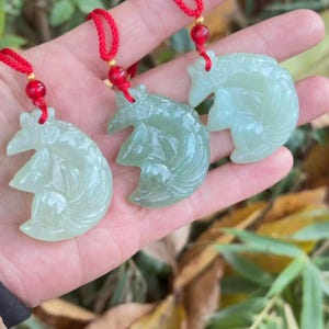 May include: Three jade pendants shaped like a stylized horse. Each pendant is a light green colour and is strung on a red cord with a gold bead and a red bead. The pendants are held in a hand.