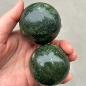 May include: Two polished, dark green spheres with a marbled appearance are held in a hand. The spheres have a smooth, reflective surface, and the color varies from deep forest green to lighter shades.