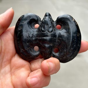 May include: A dark, possibly antique, carved stone artifact. The object is held in a hand and has a symmetrical, butterfly-like shape with rounded lobes and circular openings. The surface appears smooth and polished.