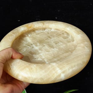 May include: A round, white, and beige alabaster dish with a smooth, slightly textured surface. The dish has a shallow, rounded well in the center.