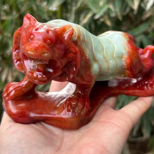 Può includere: Statuetta di tigre scolpita nei toni del rosso e del verde. La tigre è in posizione accovacciata, con la bocca aperta, che rivela i denti. La statuetta è tenuta in mano, mostrando gli intricati dettagli della scultura.