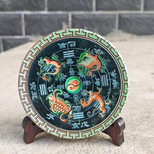 Könnte beinhalten: Runde, dunkelgrüne dekorative Scheibe mit detailliertem Design. Sie zeigt vier orange und grüne Tierfiguren, die ein zentrales Yin-Yang-Symbol umgeben. Die Scheibe wird von einem kleinen Holzständer getragen.