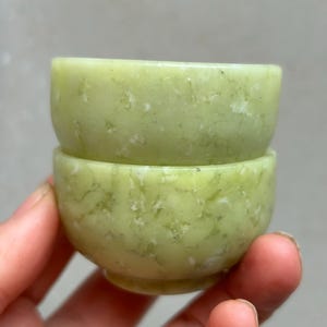 May include: Two small, light green jade bowls stacked. The bowls have a smooth, rounded shape with a slightly textured surface. The jade has natural variations in colour, with white and darker green marbling. The bowls are likely around 7 cm in diameter.