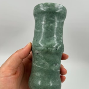 Puede incluir: Una herramienta de masaje de jade verde con forma de tallo de bambú. La herramienta tiene una superficie lisa y pulida con variaciones naturales de color. El jade es verde claro con motas verdes más oscuras y negras. Mide unos 10 cm.