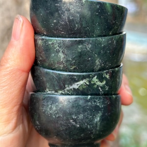 Könnte beinhalten: Ein Stapel von vier kleinen, dunkelgrünen Steinschalen. Die Schalen haben eine glatte, abgerundete Form mit einem kleinen Fuß. Der Stein weist natürliche Farbvariationen mit helleren grünen Adern auf. Jede Schale hat einen Durchmesser von etwa 6 cm.