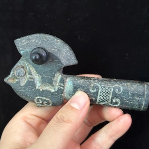 May include: A dark gray, carved stone pipe with a bird-like design. The pipe features intricate carvings and a dark, round bead. The pipe is held in a hand against a black background.