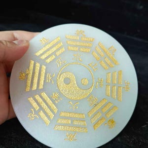 Könnte beinhalten: Ein runder, cremefarbener Stein mit goldfarbenen chinesischen Schriftzeichen und einem Yin-Yang-Symbol in der Mitte. Der Stein wird in einer Hand vor dunklem Hintergrund gehalten. Das Design ist symmetrisch und detailliert.