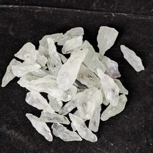 May include: A collection of clear, crystalline quartz points. The quartz crystals are translucent with a pale green hue, and vary in size and shape. They are scattered on a dark surface, creating a contrast that highlights their natural beauty.