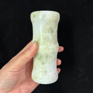 Puede incluir: Objeto cilíndrico de piedra verde claro y blanca con diseño de bambú. La piedra tiene una superficie pulida y un aspecto veteado natural. El objeto se sostiene en una mano sobre un fondo negro.