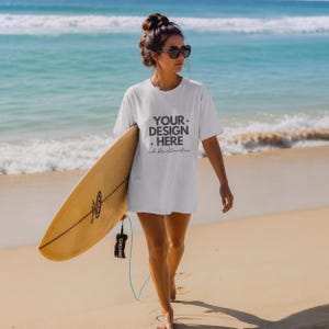 May include: A white short-sleeved t-shirt featuring the dark gray text "YOUR DESIGN HERE" with small star icons and cursive script below. The shirt is worn with dark sunglasses and is paired with a light brown surfboard held at the side.