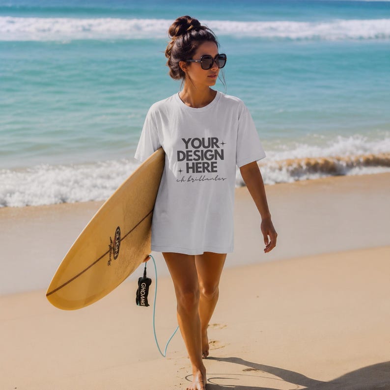 May include: A white short-sleeved t-shirt featuring the dark gray text "YOUR DESIGN HERE" with small star icons and cursive script below. The shirt is worn with dark sunglasses and is paired with a light brown surfboard held at the side.