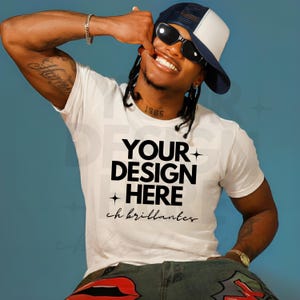 Puede incluir: Camiseta blanca con el texto "YOUR DESIGN HERE" en negrita, negro. La camiseta se usa con una gorra de béisbol azul marino y blanca y gafas de sol. El modelo sonríe y lleva un reloj y una pulsera dorados.