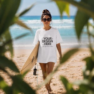 May include: A white short-sleeved t-shirt featuring the black text "YOUR DESIGN HERE" with small star icons and the cursive script "oh brillante" printed on the front. The shirt is worn with dark sunglasses while holding a light-colored surfboard.