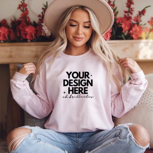 Puede incluir: Una sudadera rosa claro con mangas largas. La parte delantera de la sudadera presenta el texto "YOUR DESIGN HERE" en negrita, en negro, con las palabras "ch brillanter" debajo. La modelo lleva vaqueros azules y un sombrero beige.