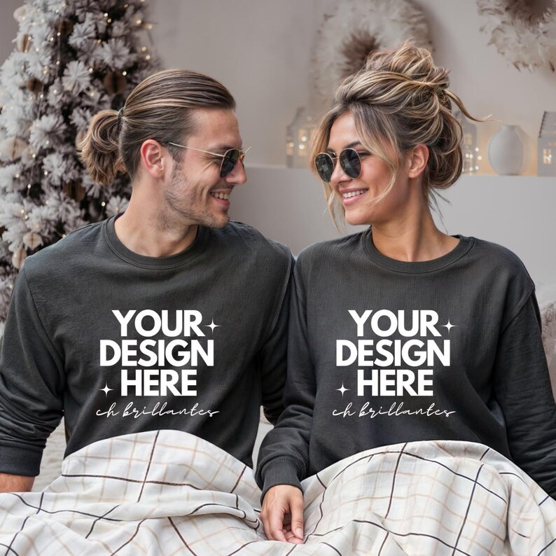 May include: Two people wearing dark gray sweatshirts with the text "YOUR DESIGN HERE" in white. The sweatshirts have long sleeves and a crew neck. The text "ch brillanter" is below the main text. They are sitting on a bed.