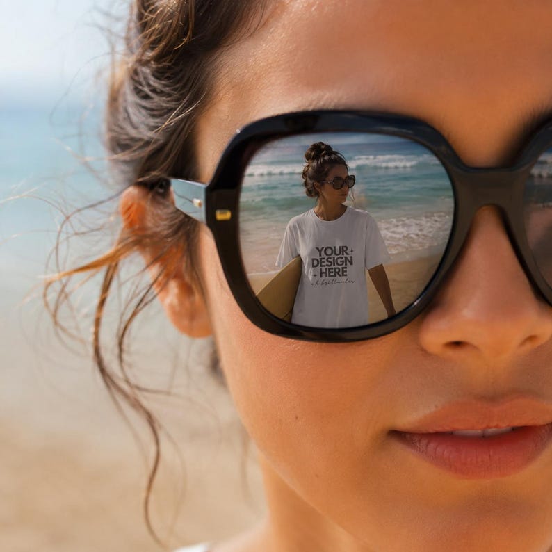 May include: Black-framed sunglasses with a reflective lens showing a person on a beach wearing a white t-shirt with the text YOUR DESIGN HERE. The reflection captures a sandy shoreline, ocean waves, and a person holding a surfboard in the background.