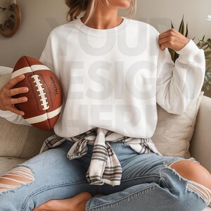 May include: A white crewneck sweatshirt is held near a brown and white football. The sweatshirt is paired with light blue ripped jeans and a plaid shirt tied around the waist. The football has white laces.