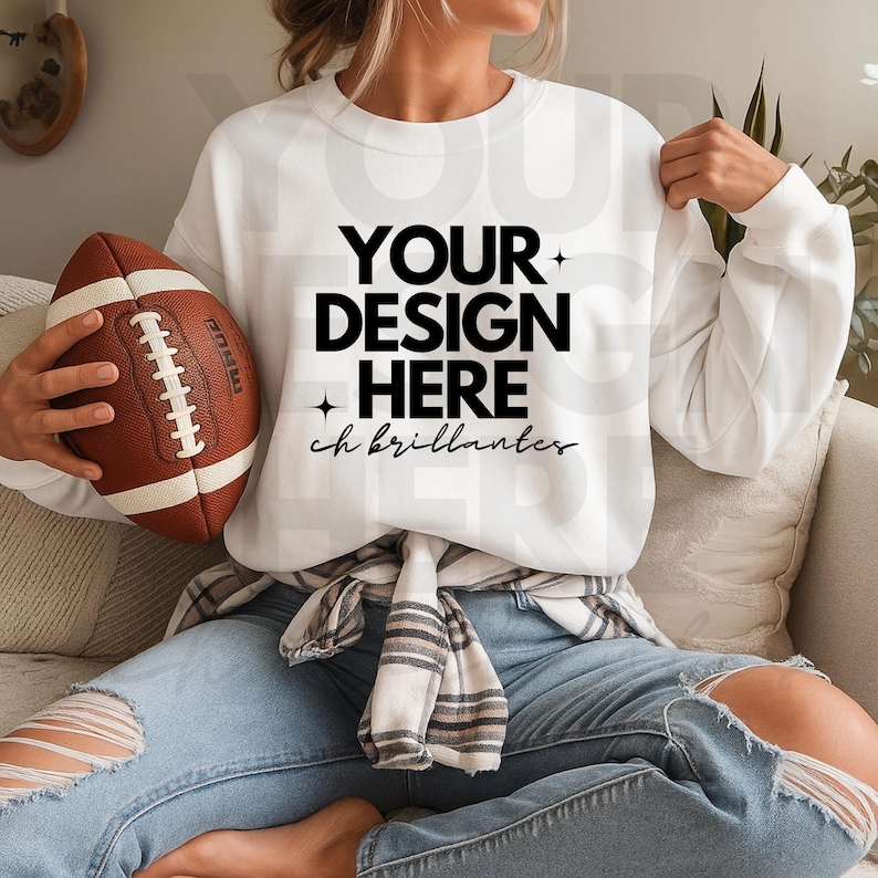 May include: A white sweatshirt with the text "YOUR DESIGN HERE" in black. A brown and white football is held in the left hand. The sweatshirt is paired with blue jeans.