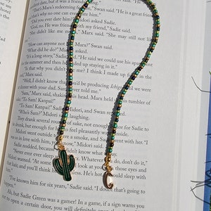 May include: A close-up of a book with text about a character named Marx. A green, gold, and black beaded necklace with a cactus and a letter C charm is lying on top of the book.