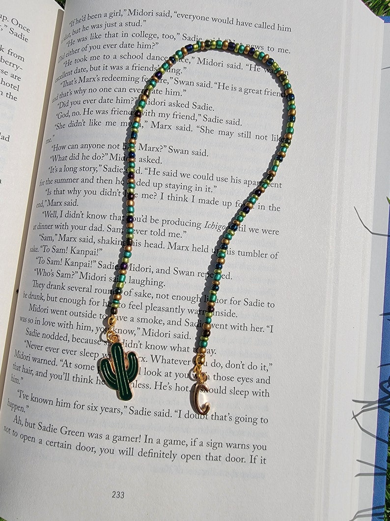 May include: A close-up of a book with text about a character named Marx. A green, gold, and black beaded necklace with a cactus and a letter C charm is lying on top of the book.