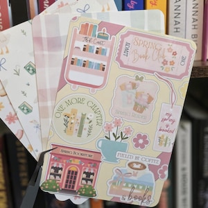 May include: A collection of decorative bookmarks with pastel colors and spring-themed illustrations. Designs include a bookstore, floral arrangements, and the text "Spring Book Club". The bookmarks are arranged in front of a bookshelf.