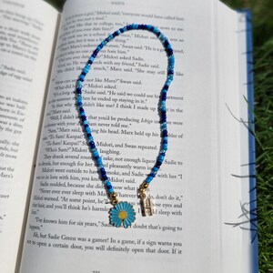 May include: A blue and white beaded necklace with a gold charm of a daisy flower. The necklace is lying on an open book with text about a character named Marx.