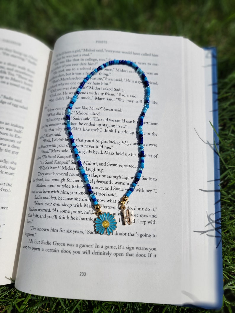 May include: A blue and white beaded necklace with a gold charm of a daisy flower. The necklace is lying on an open book with text about a character named Marx.