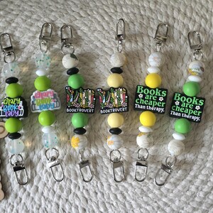 May include: Six beaded lanyards featuring silver lobster clasps and silicone beads in shades of green, yellow, and white. The charms include text reading "crazy book lady," "BOOKTROVERT," and "Books are cheaper than therapy." A wooden sign reads "readingwiththreeses."