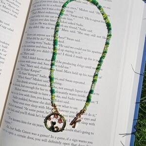 May include: A green and white beaded necklace with a charm of a cat sitting in a crescent moon. The charm is gold and has a green enamel finish. The necklace is lying on a book with the text "PIVOTS" visible.