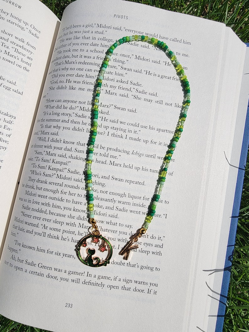 May include: A green and white beaded necklace with a charm of a cat sitting in a crescent moon. The charm is gold and has a green enamel finish. The necklace is lying on a book with the text "PIVOTS" visible.