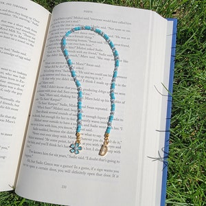 May include: A turquoise and silver beaded necklace with a gold charm and a blue flower charm. The necklace is lying on an open book with text about a character named Marx.