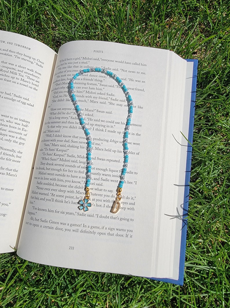 May include: A turquoise and silver beaded necklace with a gold charm and a blue flower charm. The necklace is lying on an open book with text about a character named Marx.