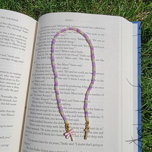May include: A close-up of a book with a purple, white, and yellow beaded necklace with butterfly charms. The book is open to a page with the text "PIVOTS" and the text "I doubt that's going to happen."