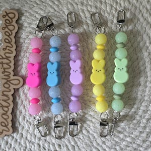 May include: Five beaded keychains in pink, blue, purple, yellow, and green. Each keychain features a Peep-shaped charm and a metal clip. The keychains are strung with various sized beads.