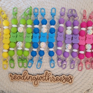 May include: Seven colorful beaded lanyards with clips on each end. The beads are shaped like a smiling marshmallow and are in the colors yellow, green, blue, purple, and pink. The text "readingwithreeses" is written on a wooden sign in the bottom center of the image.