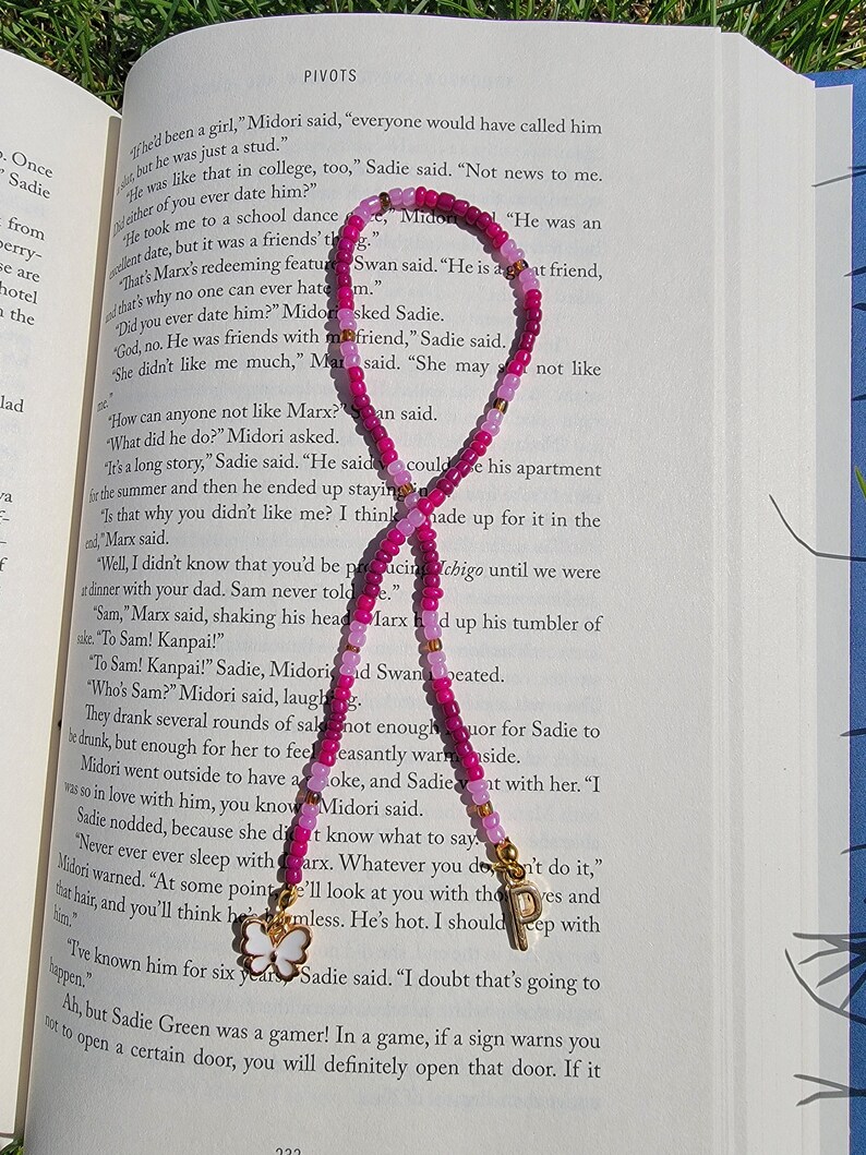 May include: Two pink and white beaded bracelets with gold butterfly charms and a gold letter "P" charm. The bracelets are on a page of a book with text about a character named Marx.