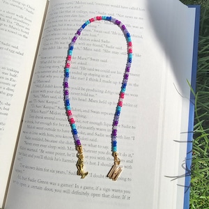 May include: A colourful beaded necklace with blue, pink, purple and white beads. The necklace has gold letter charms that spell out "M" and "S". The necklace is lying on an open book with text about a character named Marx.