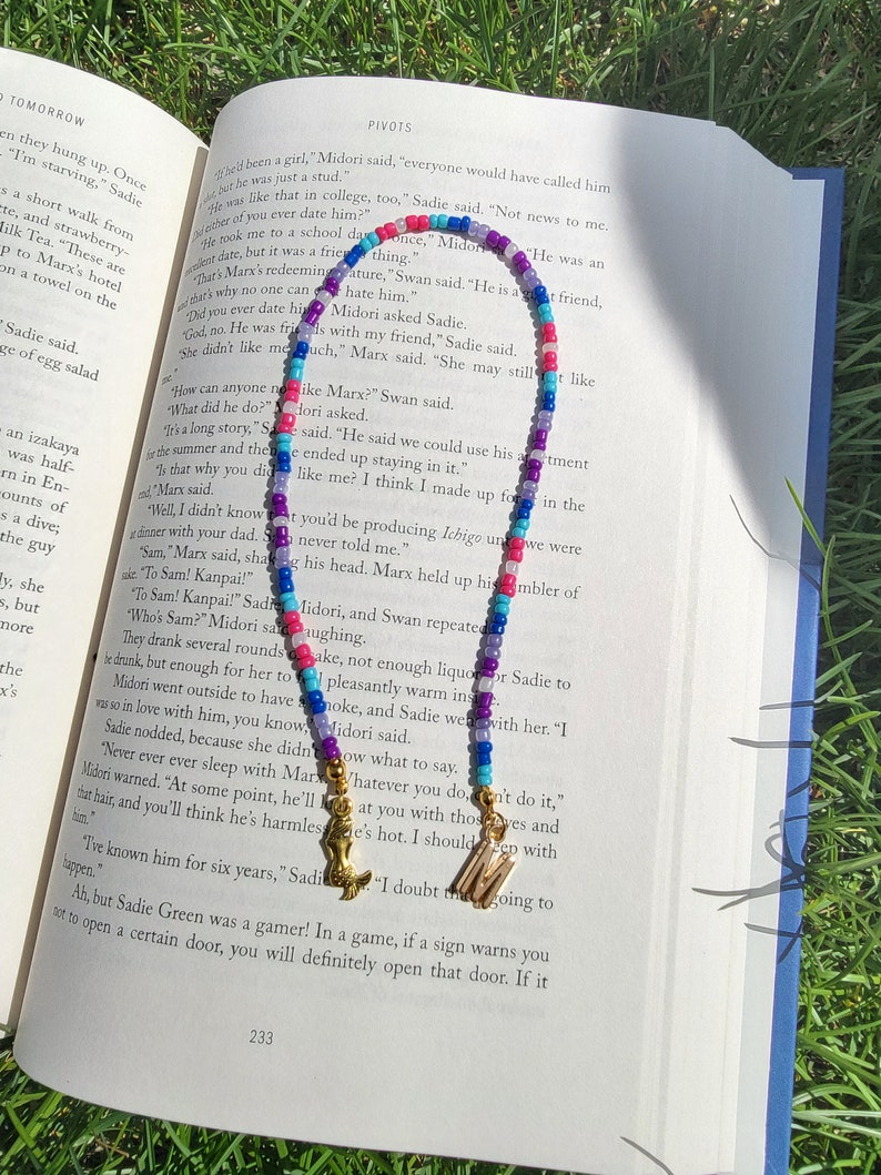 May include: A colourful beaded necklace with blue, pink, purple and white beads. The necklace has gold letter charms that spell out "M" and "S". The necklace is lying on an open book with text about a character named Marx.