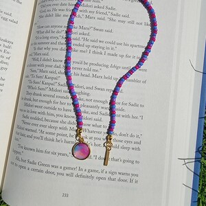 May include: A pink, blue, and purple beaded necklace with a gold charm featuring a watercolor-style design. The necklace is lying on a book with text about a character named Sadie.