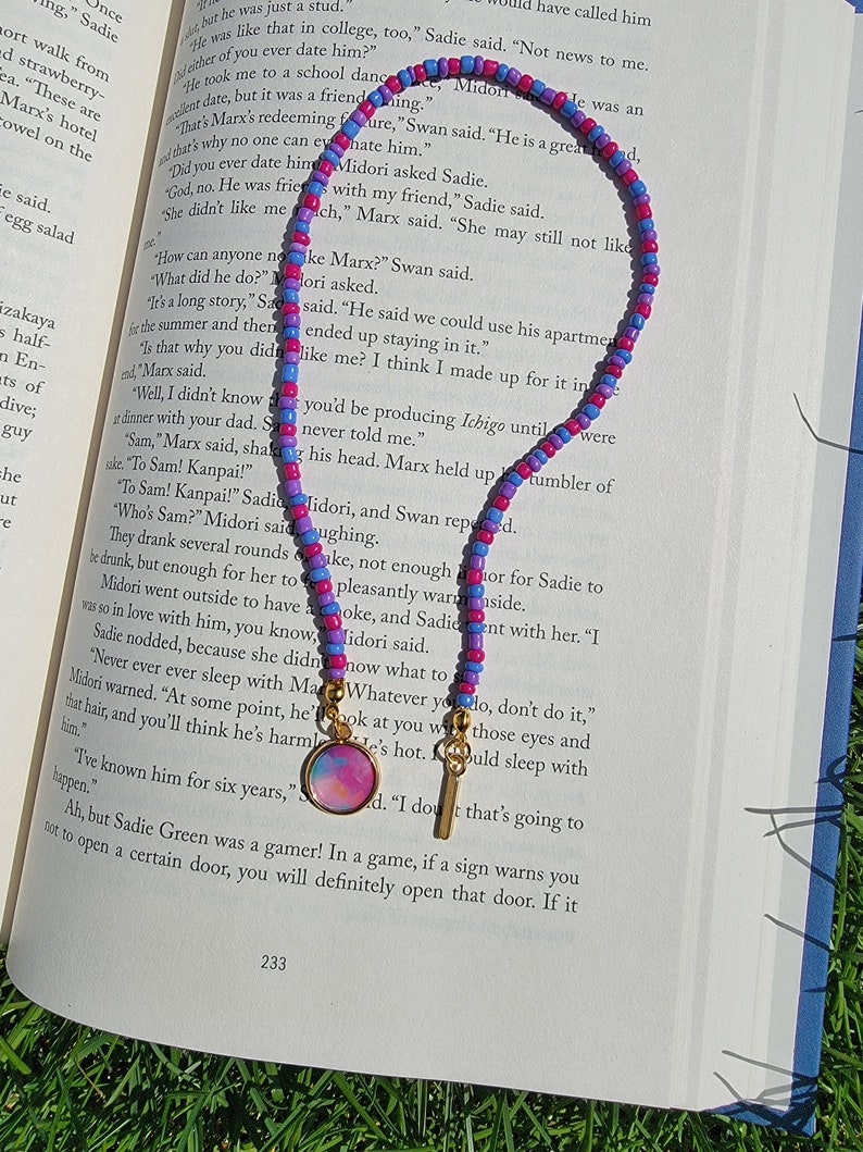 May include: A pink, blue, and purple beaded necklace with a gold charm featuring a watercolor-style design. The necklace is lying on a book with text about a character named Sadie.