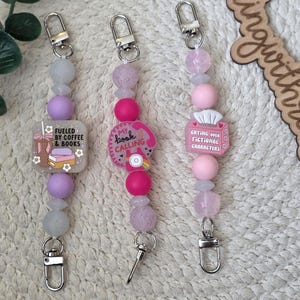 May include: Three beaded keychains with silver clasps. One keychain has a square charm with the text "Fueled by Coffee & Books." Another has a pink charm that says "My Book Calling." The third keychain has a charm that says "Crying Over Fictional Characters."