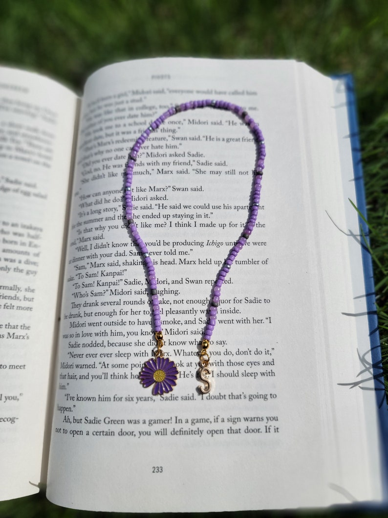 May include: A purple beaded bracelet with a gold charm featuring a flower and the letter "S". The bracelet is lying on a book with text about a character named Marx.