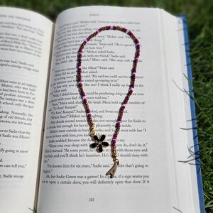 May include: A close-up of a book with a beaded bookmark featuring a black flower charm. The bookmark is purple and gold with white accents. The book is open to a page with text about a character named Sadie.