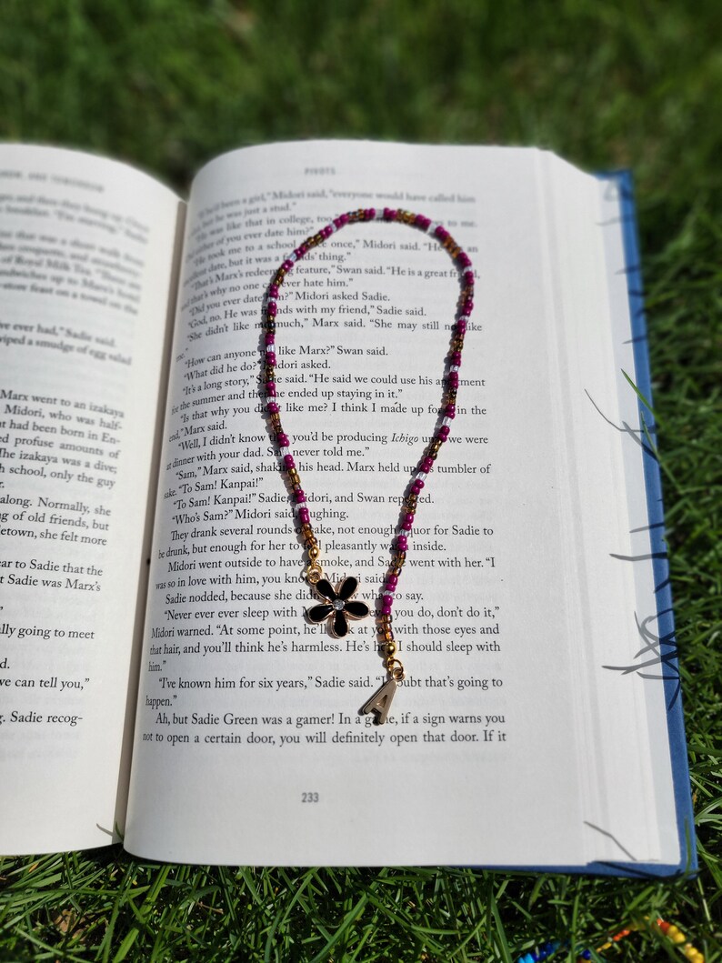 May include: A close-up of a book with a beaded bookmark featuring a black flower charm. The bookmark is purple and gold with white accents. The book is open to a page with text about a character named Sadie.